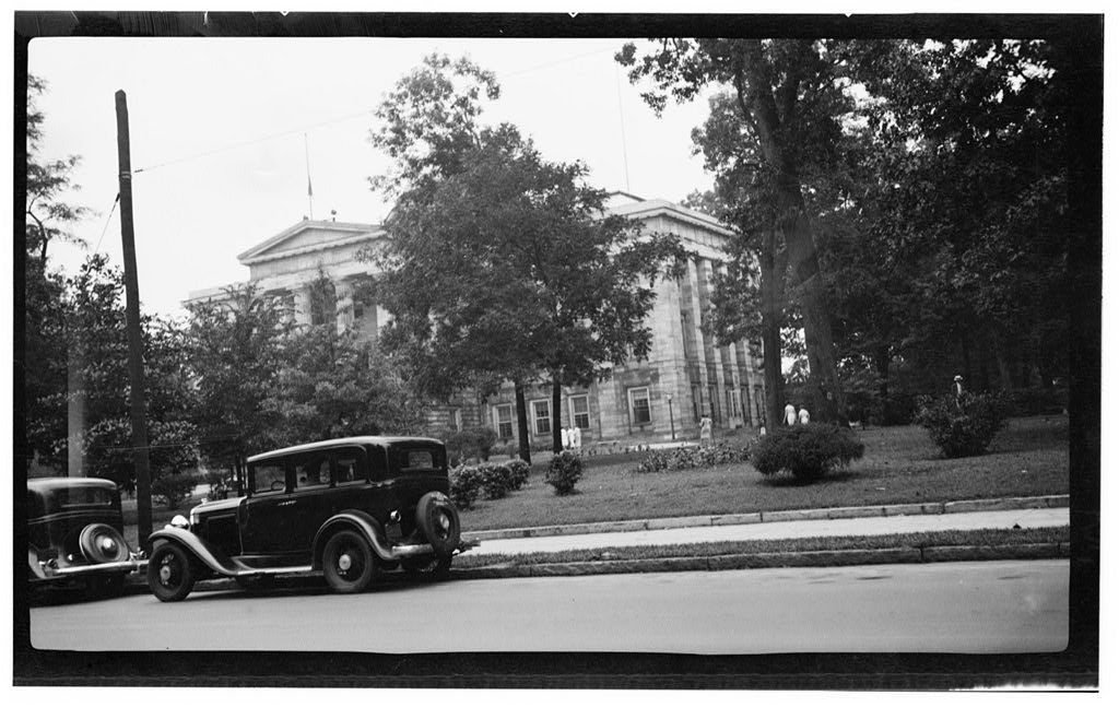 Stone North Carolina State Capitol behind trees on Capitol Square, with 1930s cars on the street
