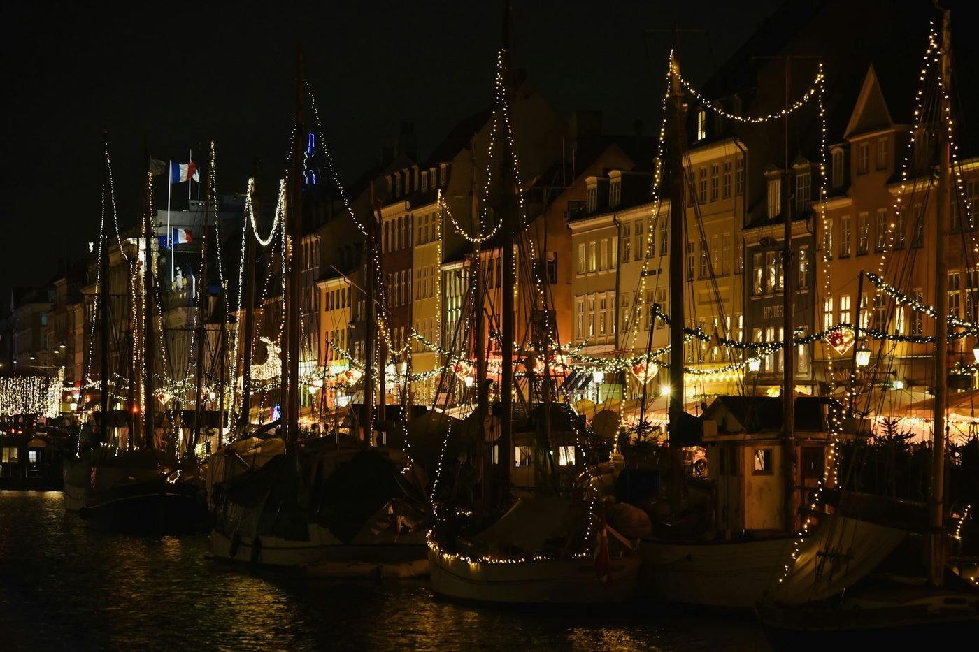Sailboats outlined in Christmas lights beside waterfront buildings at night (illustrative photo by Danny Chen on Unsplash, not the Wrightsville Beach Flotilla)